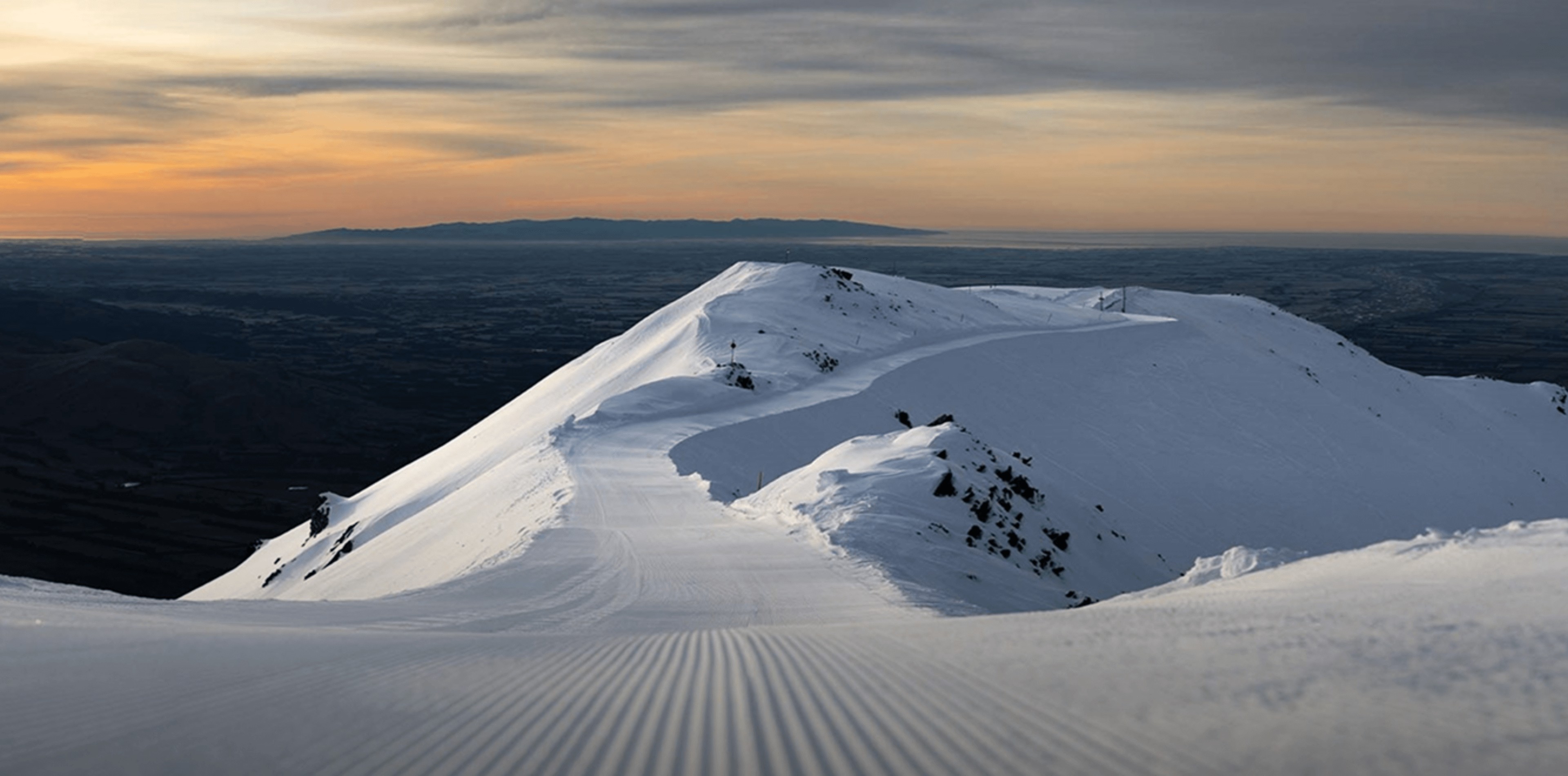 Snow-covered mountain ridge with a groomed ski trail at sunset.