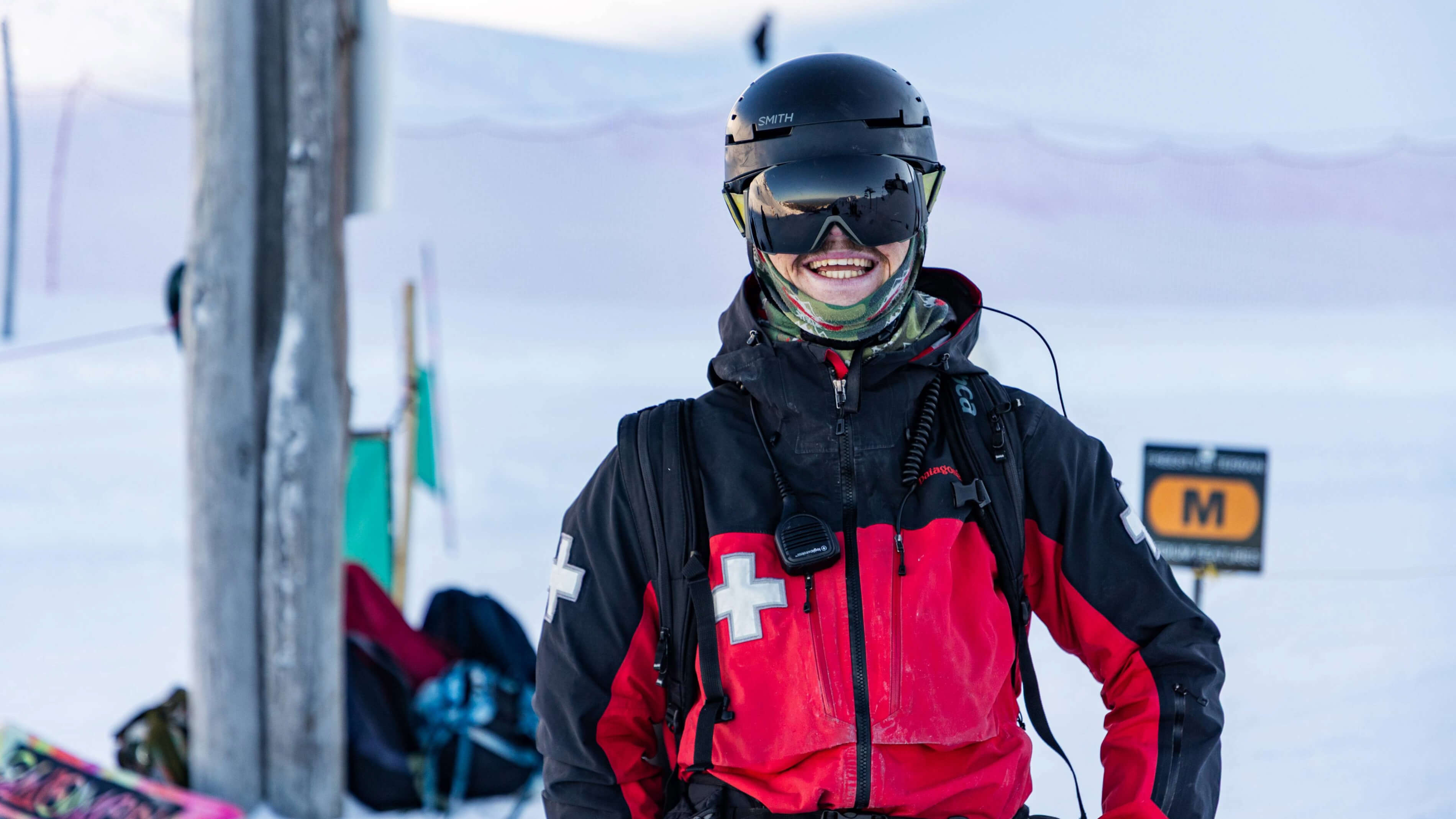 Smiling ski patroller in red and black jacket wearing helmet and goggles in snowy environment.