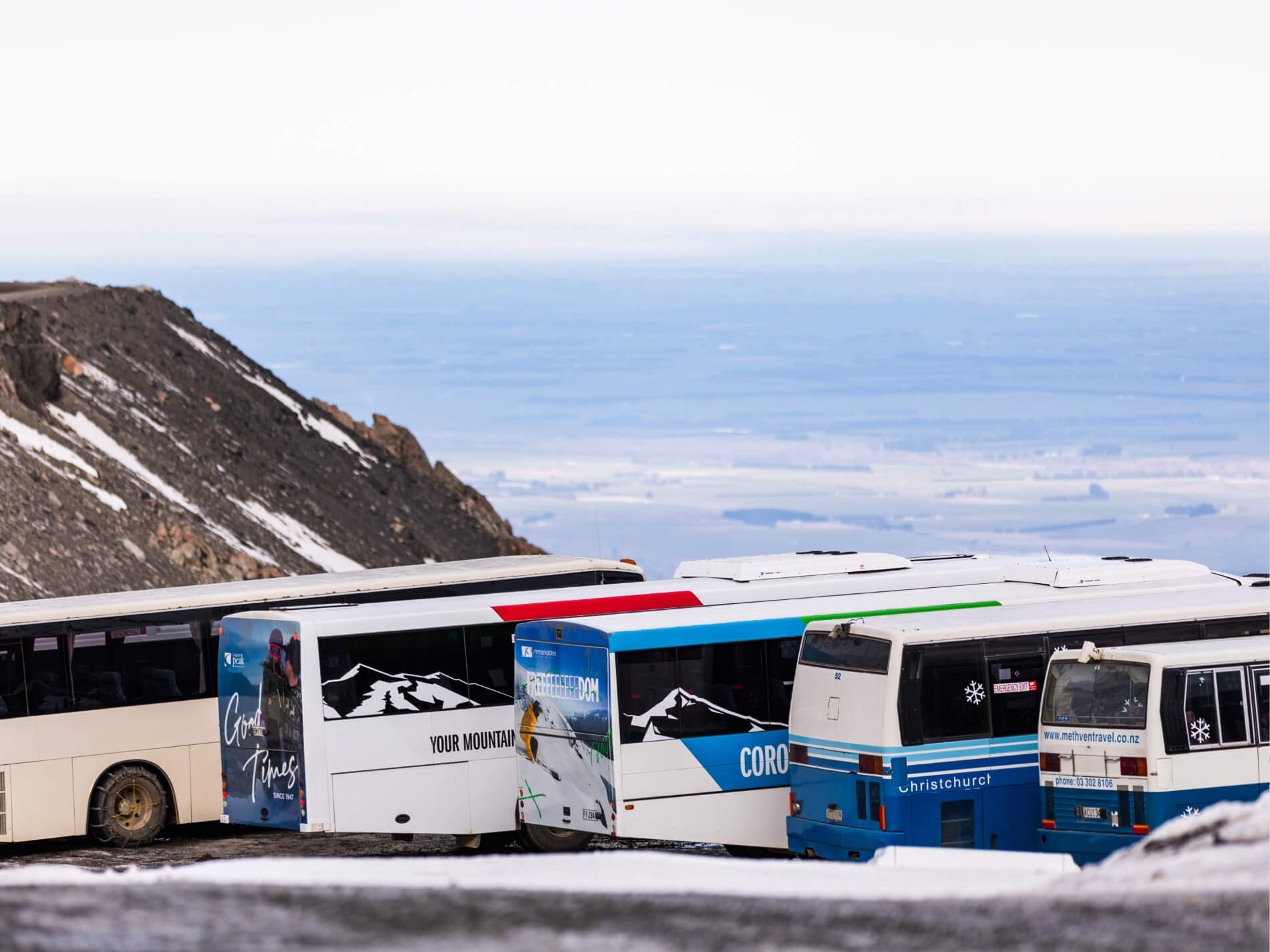 Multiple parked tour buses on a snowy mountain road with a distant valley visible in the background.
