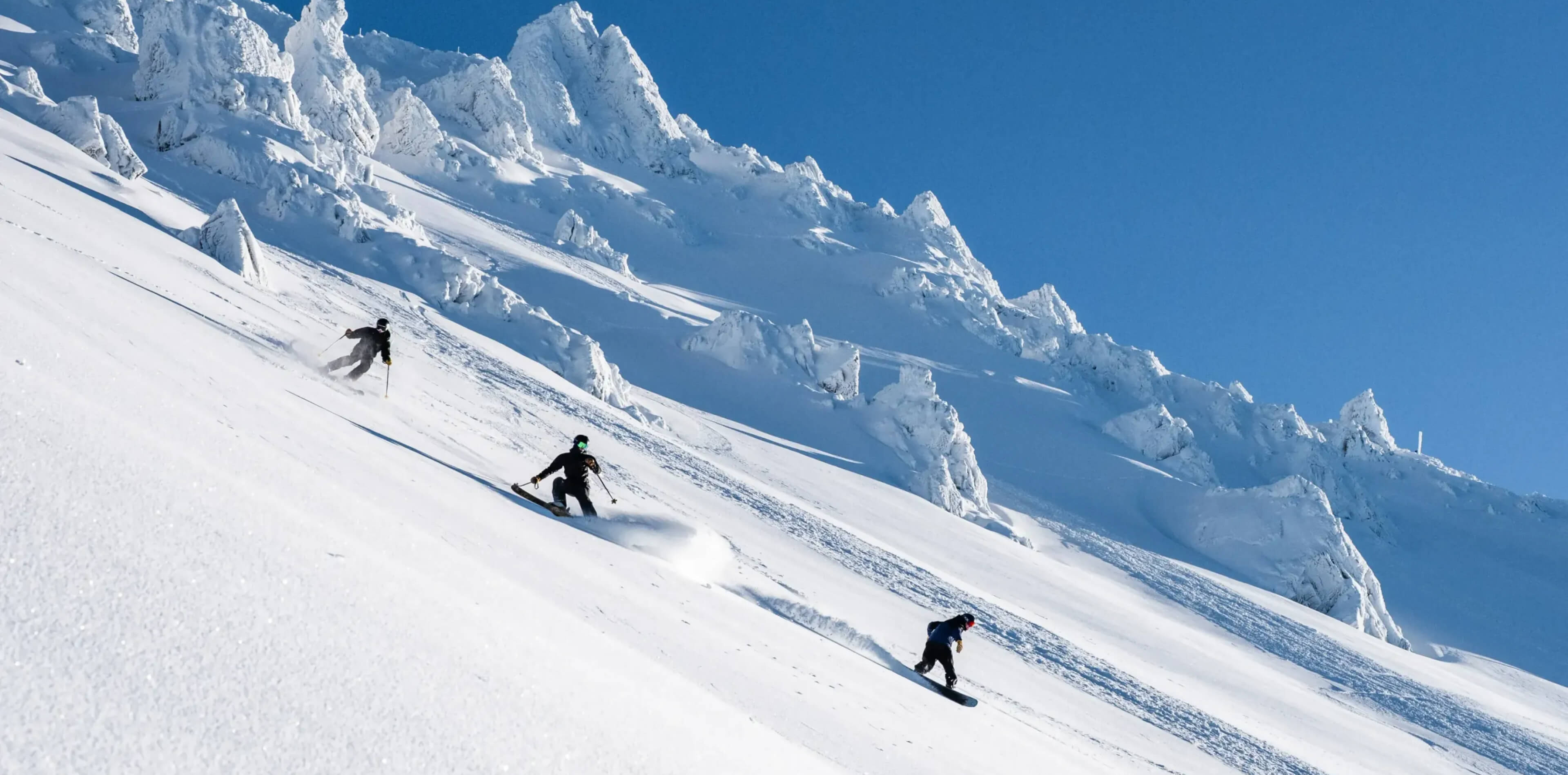 Three people skiing and snowboarding down a snow-covered mountain slope at the top of Mt Hutt under a clear blue sky.